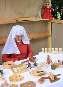 Frau in hochmittelalterlicher Bekleidung vor Tisch mit Spielzeug. Sie trägt ein rotes Kleid und ein weißes Gebende auf dem Kopf. Im Hintergrund eine Stoffpuppe zum spielen, die ähnlich wie die Frau berkleidet ist.